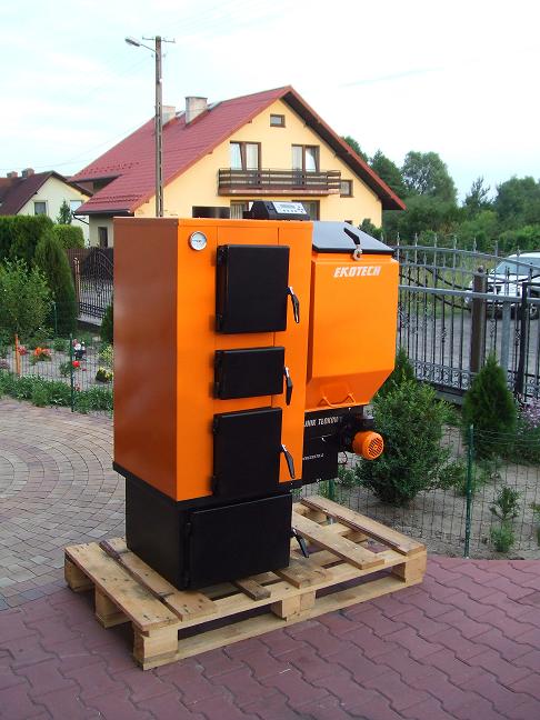 Pomarańczowo-czarny piec z podajnikiem tłokowym EKON, umieszczony na drewnianej palecie, na tle domu jednorodzinnego i ogrodzenia.